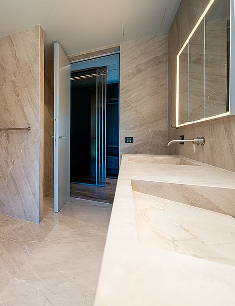 Modern master bathroom featuring a white oval freestanding bathtub against beige stone walls, a double vanity, and recessed LED lighting.