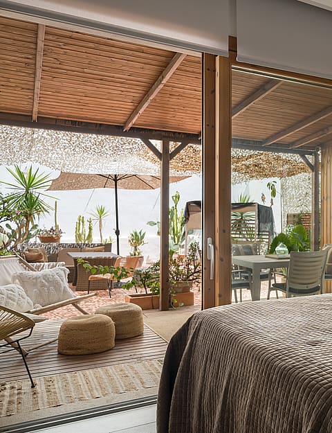 A bedroom with a textured brown bedspread, opening through large sliding glass doors to a furnished plant-filled terrace.