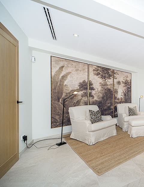 Interior room featuring beige armchairs on a jute rug, a large forest-motif wall mural, and light stone flooring.