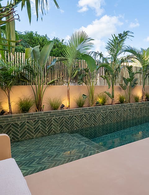 A narrow swimming pool with green patterned tiles is surrounded by a beige stone terrace and lush palm trees.