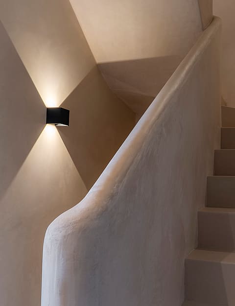 A staircase with off-white plaster walls and a textured balustrade, illuminated by a sharp geometric wall light.