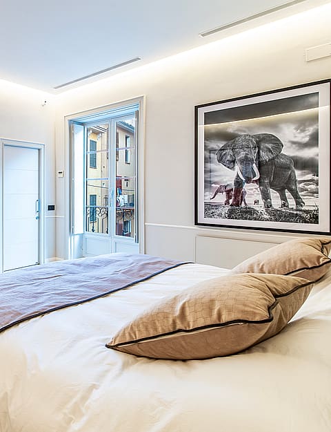 A modern bedroom featuring a king-sized bed, cream-colored armchair, recessed cove lighting, and a view of an Old Town street.