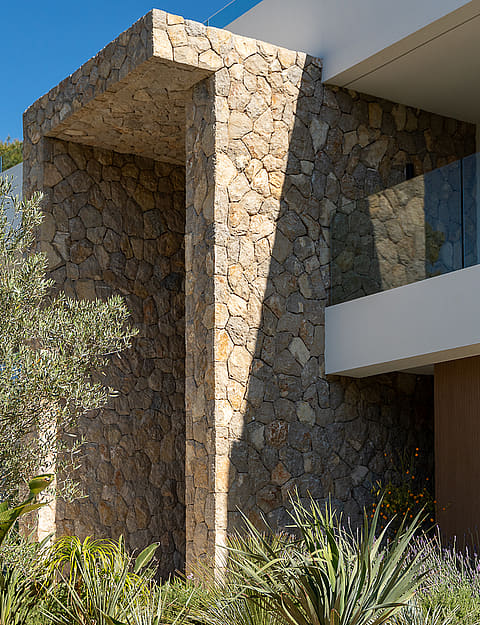 Close up of a modern villa exterior showing dry stone wall architecture, an olive tree, and a glass balcony railing.