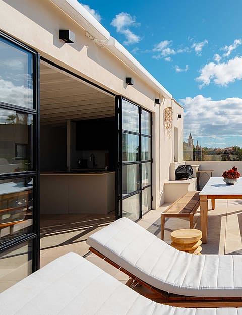 A sunny rooftop terrace in Santa Catalina with white sun loungers, a wooden dining table and benches, and iron-framed glass doors.