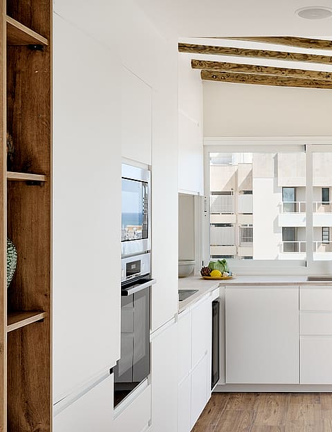 Modern kitchen with white cabinets, wood ceiling beams, and a window overlooking an urban exterior.