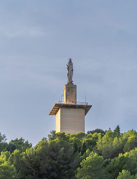 A stone statue on a pedestal positioned atop a lush green hill against a clear sky in Genova.