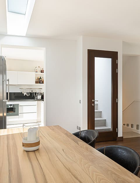 Bright dining space with large oak table, stainless steel fridge, and a skylight in a modern white interior.