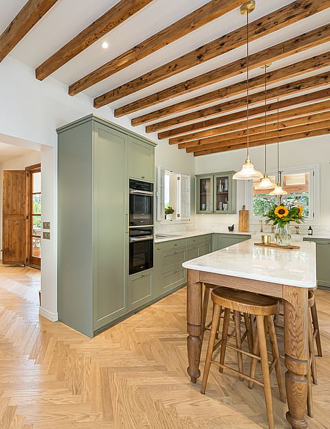 Modern kitchen featuring sage green cabinets, herringbone wood floors, a marble-top island, and exposed timber ceiling beams.