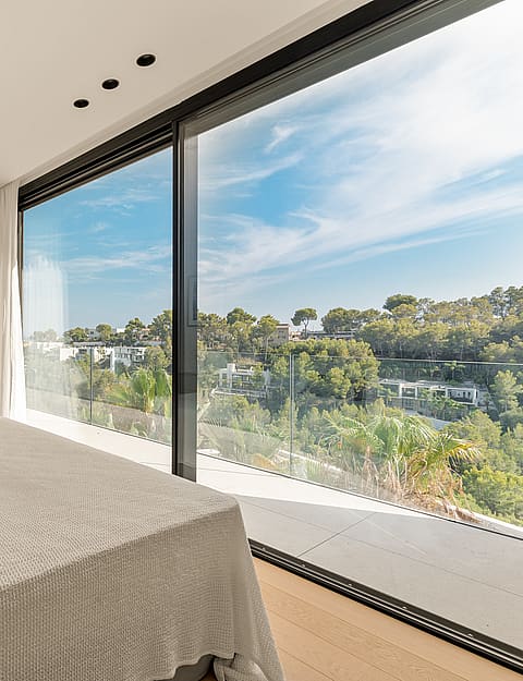 A modern bedroom with a large bed, featuring floor-to-ceiling sliding glass doors that open to a green landscape view.