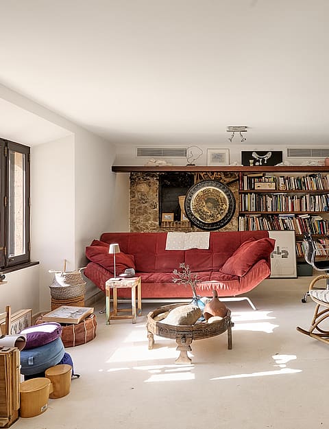 A living area in a Finca featuring a large stone fireplace, exposed wooden beams, a red sofa, and a wicker rocking chair.