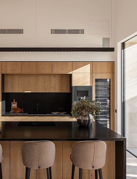A modern kitchen with wood cabinetry, a black stone island, and three upholstered stools, opening to a sunlit terrace.