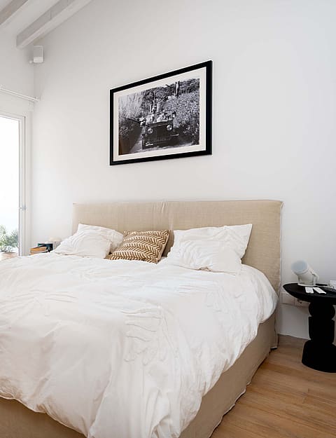Modern bedroom with a neutral upholstered bed, light oak wood flooring, white walls, and a door leading to a bathroom.