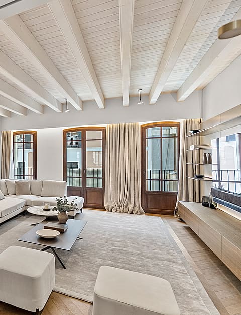 Bright living room with herringbone wood flooring, white sectional sofa, and tall wooden French doors under a beamed ceiling.