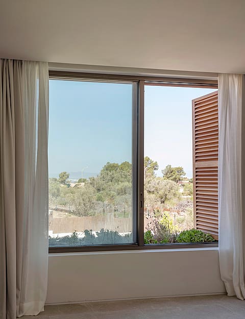 Interior view of a window with brown wooden shutters and white curtains looking out to a sun-drenched green landscape.