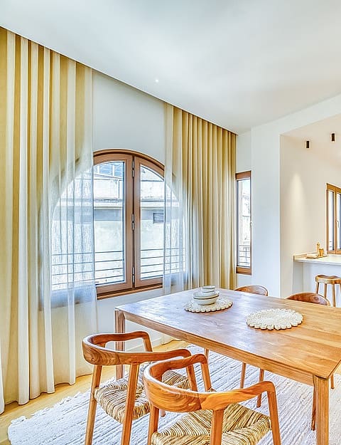 A bright open-plan kitchen and dining room with wooden furniture, white cabinetry, and large arched windows draped in curtains.