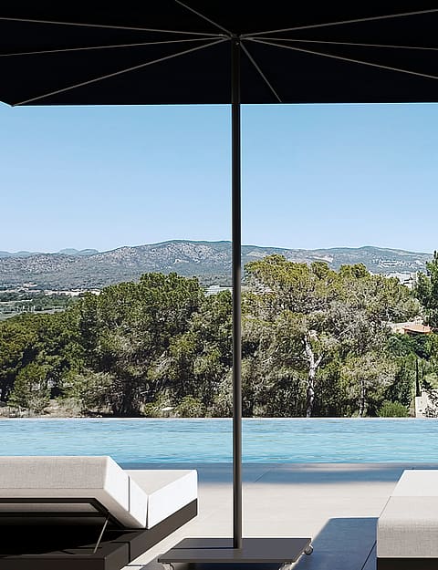 A poolside terrace with white sun loungers, a black parasol, and a clear view of pine trees and distant mountains.