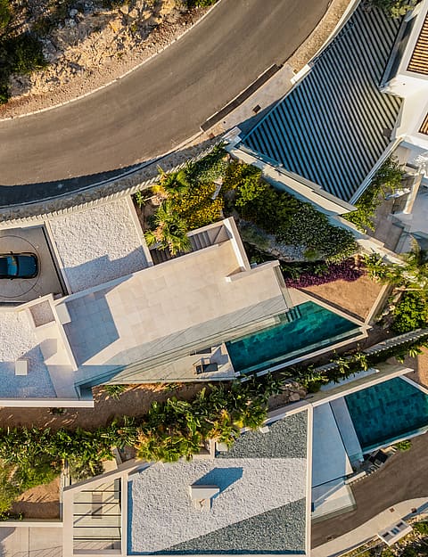 Top-down aerial view of a multi-level hillside villa showing rooftops, two swimming pools, stone terraces, and access road.