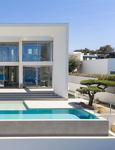 Two-story white modern villa with floor-to-ceiling glass walls, a rectangular swimming pool, and stone-paved terrace.