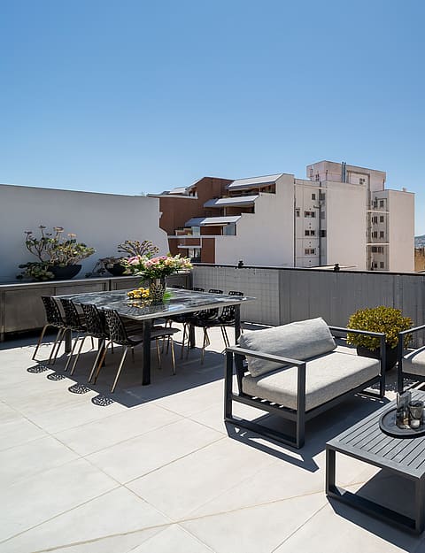 A spacious rooftop terrace featuring grey modular outdoor furniture, a glass dining table, and wide views of the city skyline.