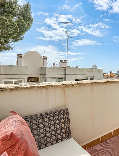 A private outdoor terrace with tiled flooring, a low perimeter wall, and patterned cushions under a bright blue sky.