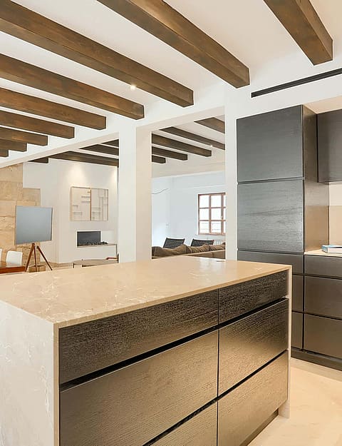 A modern kitchen with a dark central island and a dining table in an open-plan room featuring exposed ceiling beams and limestone walls.