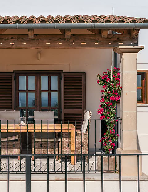 Porche con columnas de piedra, mesa de comedor de madera, contraventanas oscuras y buganvillas en flor.
