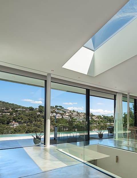 Modern living and dining room featuring floor-to-ceiling glass walls, a large skylight, and views of Son Vida hills.