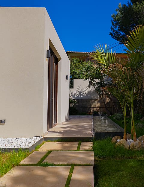 A minimalist white exterior wall with a dark framed doorway and a stone slab pathway leading through a green garden.