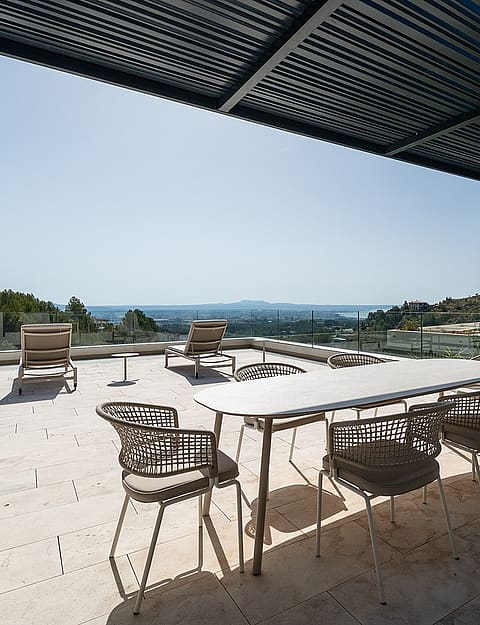 A wide outdoor terrace featuring a white stone floor, a dining table with woven chairs, and a black slatted pergola roof.