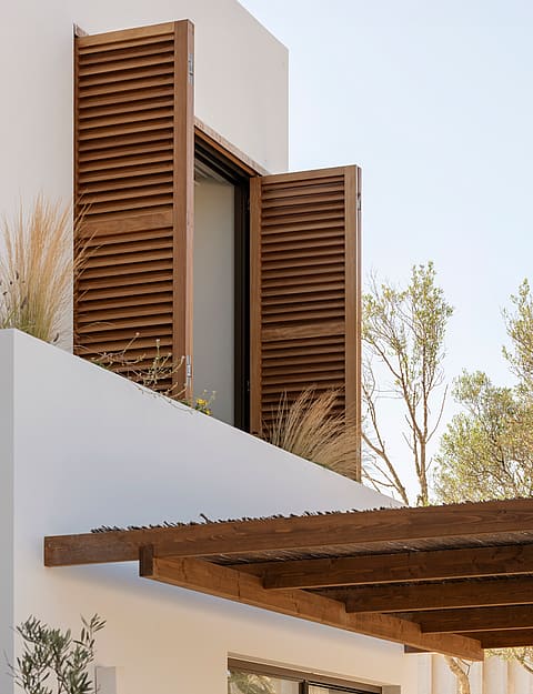 Exterior detail of a white building featuring dark wood shutters and a rustic timber pergola against a clear sky.