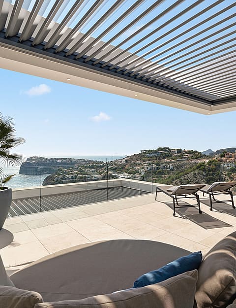 A covered terrace with a louvered pergola and lounge seating overlooking the sea and hillside villas in Port d'Andratx.