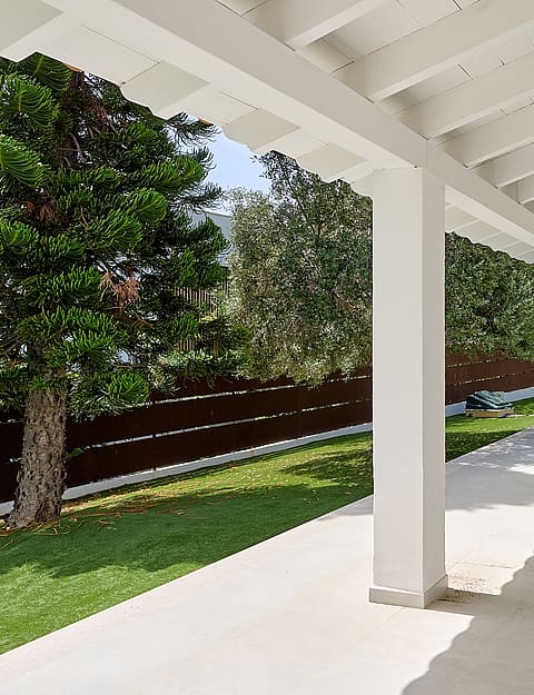 Terraza cubierta blanca con vigas de madera frente a césped artificial, piscina circular, pinos y cipreses