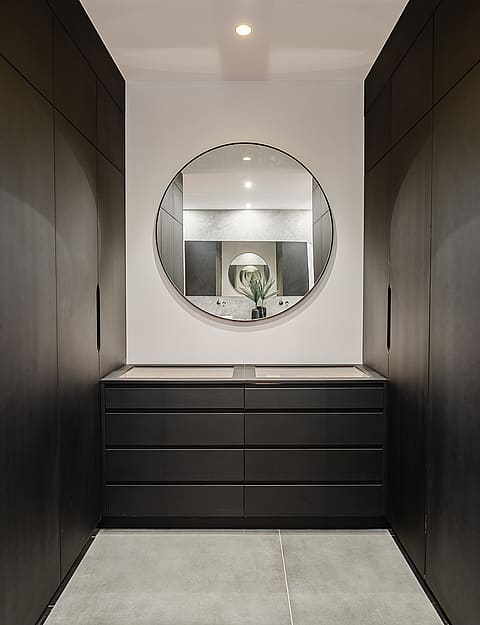 Symmetrical walk-in closet featuring floor-to-ceiling matte black cabinets, a central vanity drawer unit, and a round mirror.