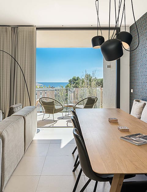 Interior view of an open-plan living and dining room with floor-to-ceiling glass doors opening onto a terrace with a sea view.