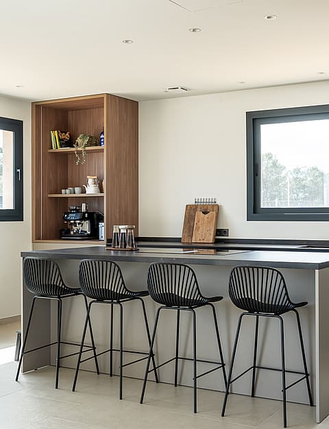 A modern kitchen with a grey island, black stools, and wooden open shelving, illuminated by natural light from windows.