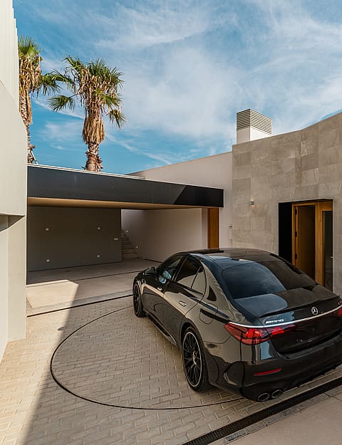 Modern outdoor courtyard featuring a stone-paved turntable for a car, shaded carport, and smooth beige walls under a blue sky.