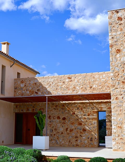 Exterior view of a stone and plaster finca featuring a terrace area with a wooden pergola, potted plants, and clear blue sky.