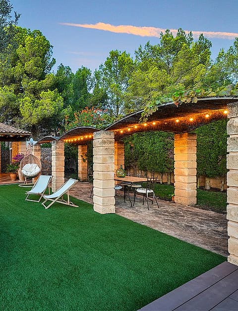 A landscaped garden with a large ancient olive tree, stone-pillared pergola with string lights, and a shaded outdoor room.