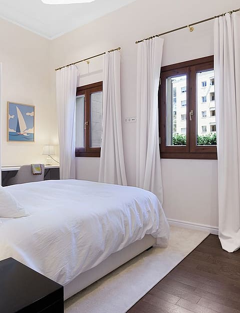 A minimalist bedroom featuring dark hardwood flooring, white walls, white bedding, and two windows with sheer curtains.