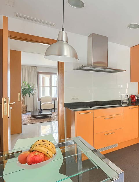 An L-shaped kitchen featuring bright orange flat-panel cabinets, black stone countertops, and a glass-topped dining table.
