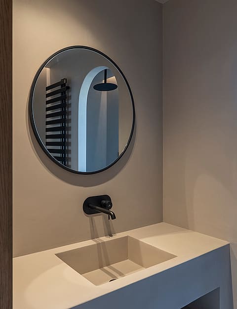 A minimalist integrated stone sink and wall-mounted black faucet beneath a circular mirror reflecting a shower area.
