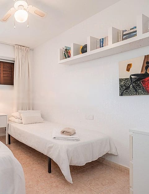 A bright guest bedroom featuring two single beds, light-toned textiles, a white wall-mounted shelf, and a minimalist aesthetic.