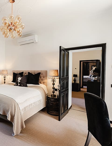 A spacious bedroom featuring a king-sized bed with dark accent pillows, a woven armchair, and an open doorway leading to another room, illuminated by a chandelier.