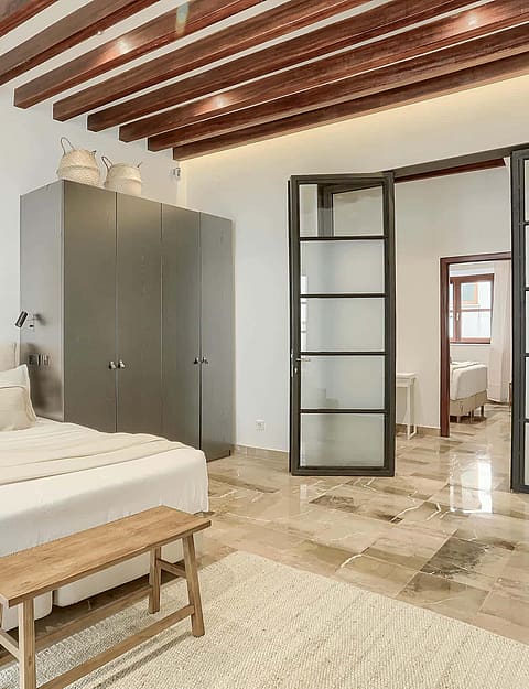 A spacious bedroom featuring exposed dark timber ceiling beams, white walls, stone flooring, and dark metal-framed glass doors.