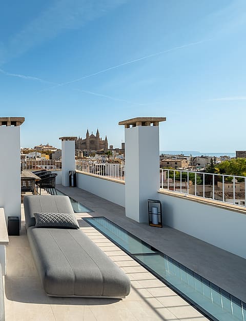 Sun-drenched rooftop terrace with grey loungers, stone flooring, and panoramic views of Palma's historic skyline.