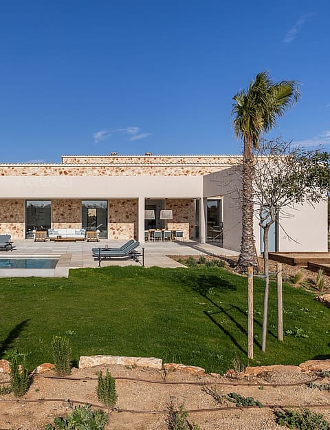 Exterior view of a modern stone finca in Santanyí featuring a rectangular swimming pool, manicured lawn, and sun loungers.