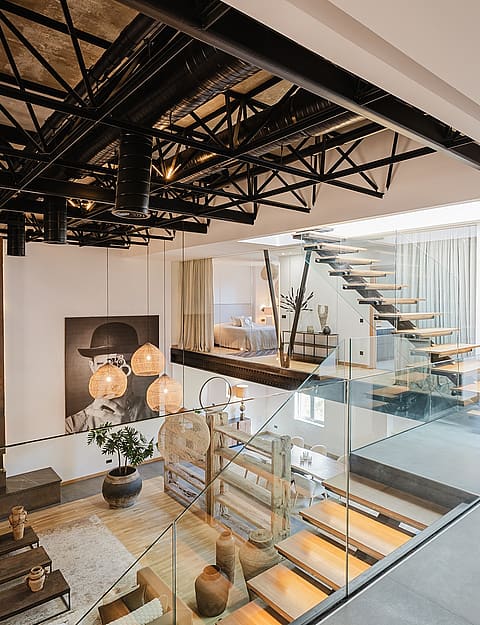High-angle view of an open-plan apartment featuring a black exposed ceiling truss, glass-railed staircase, and mezzanine level.