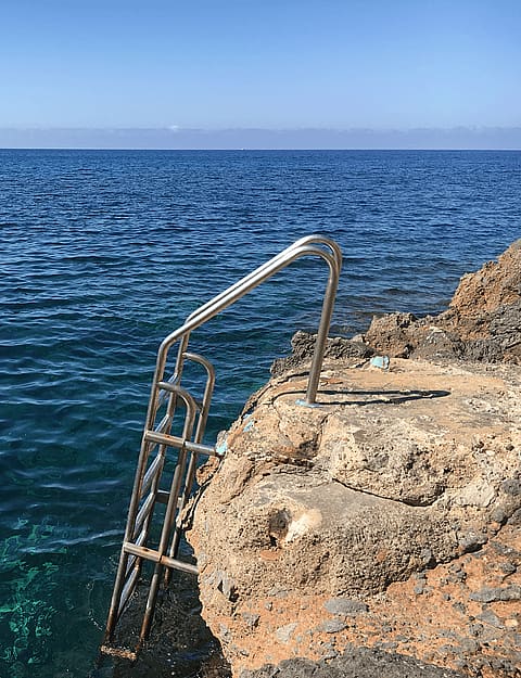 A stainless steel swim ladder attached to natural rocky coastline leading into deep blue Mediterranean sea waters.