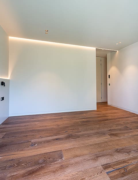 An unfurnished minimalist bedroom featuring white walls, warm LED cove lighting, and wide-plank oak wood floors.