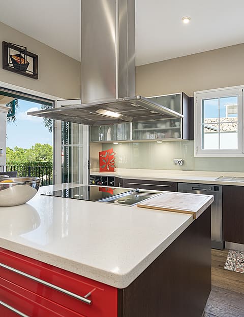 A modern kitchen featuring a bold red central island with a white stone countertop, dark cabinetry, and floor-to-ceiling glass doors opening to a terrace.